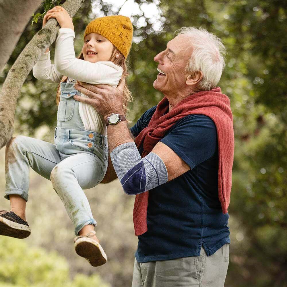 Ein älterer Mann hält ein kleines Mädchen, welche einen Ast hinaufklettert mit seinen Händen fest. Er trägt eine EpiTrain Ellenbogenbandage von Bauerfeind. Die EpiTrain Bandage lindert Schmerzen im Ellenbogen und verbessert seine Beweglichkeit.