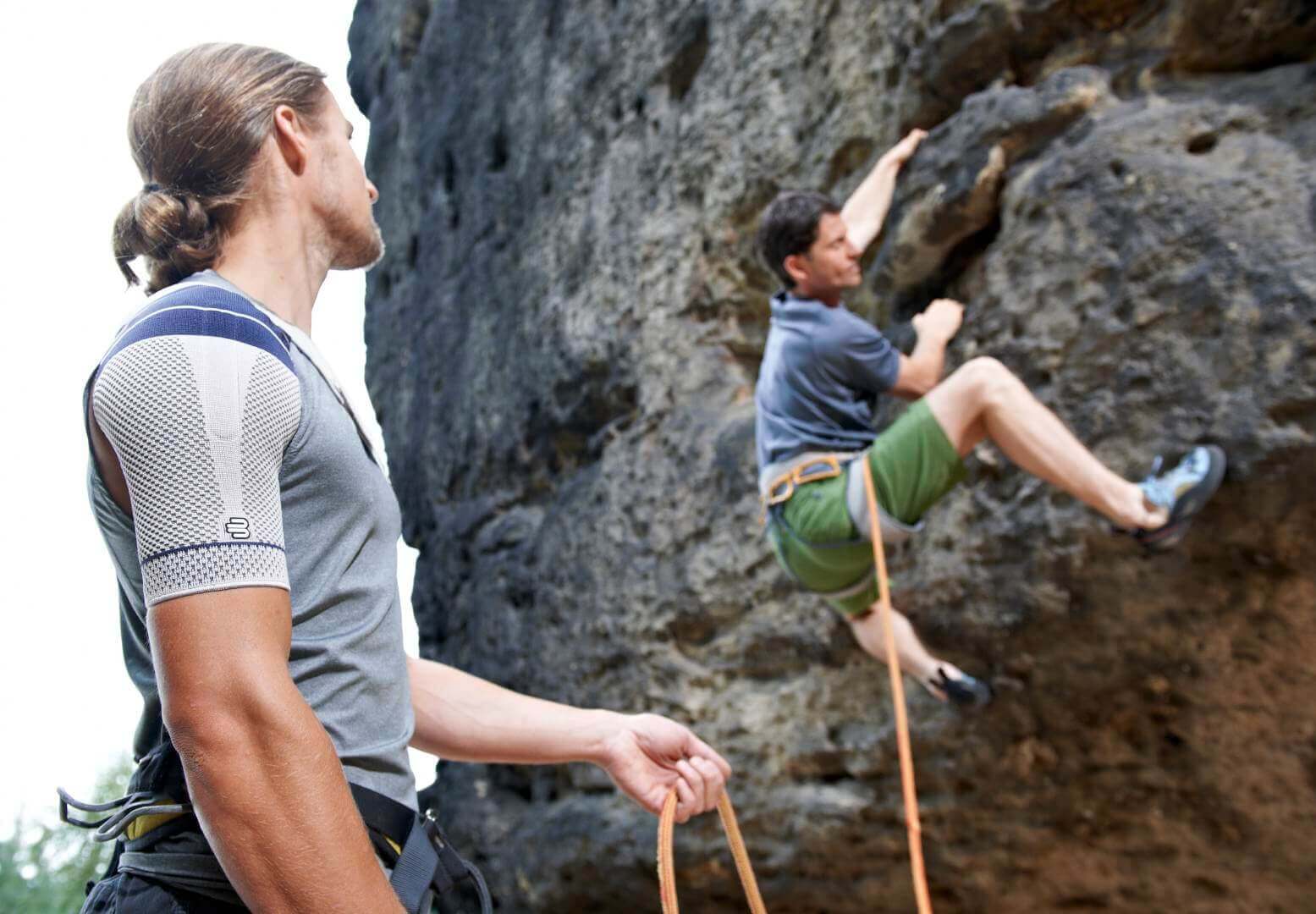 Abgebildet ist ein Duo von Kletterern, einer am Felsen, der andere sichert seinen Sportsfreund und trägt dabei eine Schulterbandage. Die OmoTrain von Bauerfeind bietet Stabilisierung und Unterstützung für das Schultergelenk.