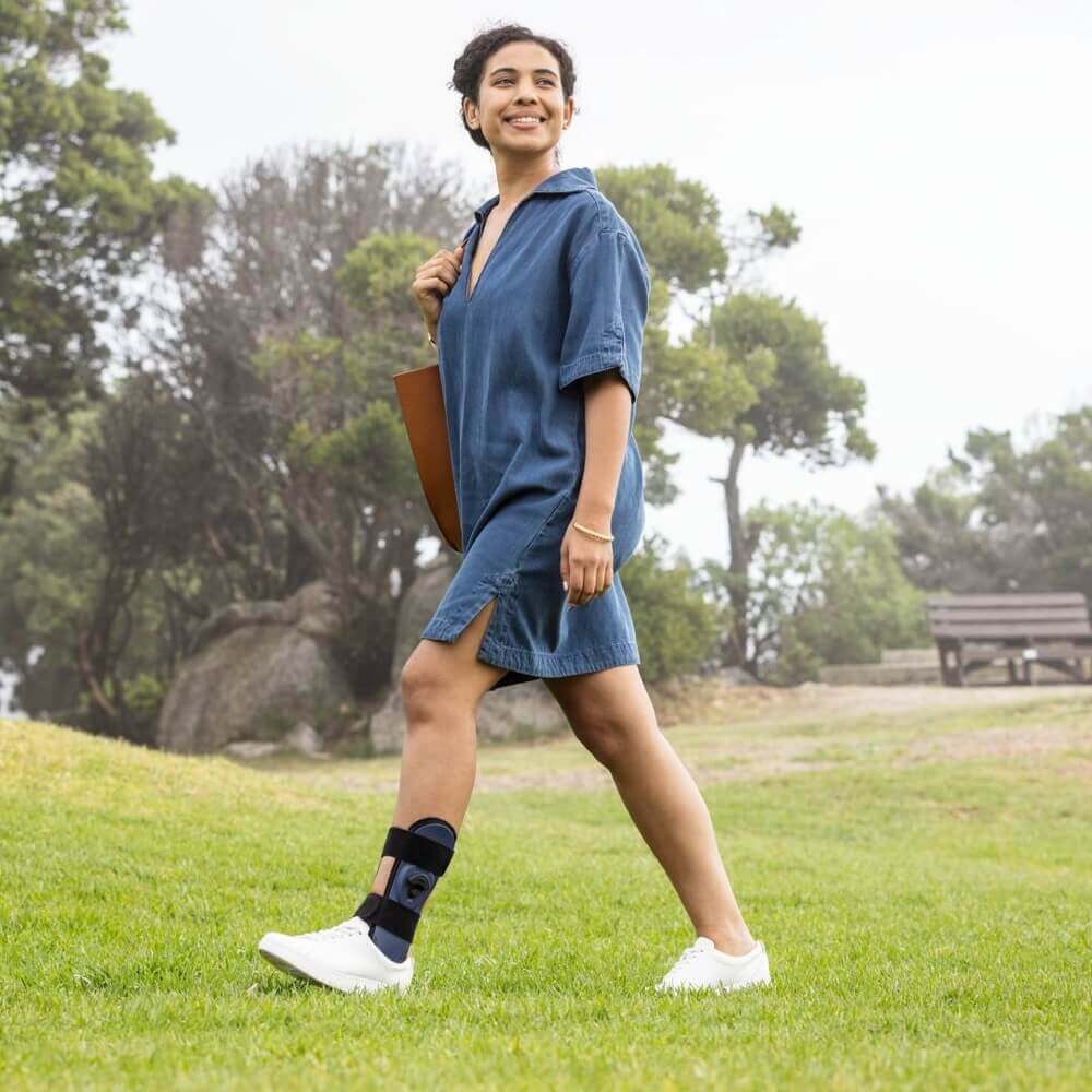 Eine junge Frau spaziert lächelnd über eine Wiese in einem Park. Sie trägt ein lockeres, kurzes Jeanskleid, weisse Turnschuhe und eine schwarze Fussorthese am linken Bein.
