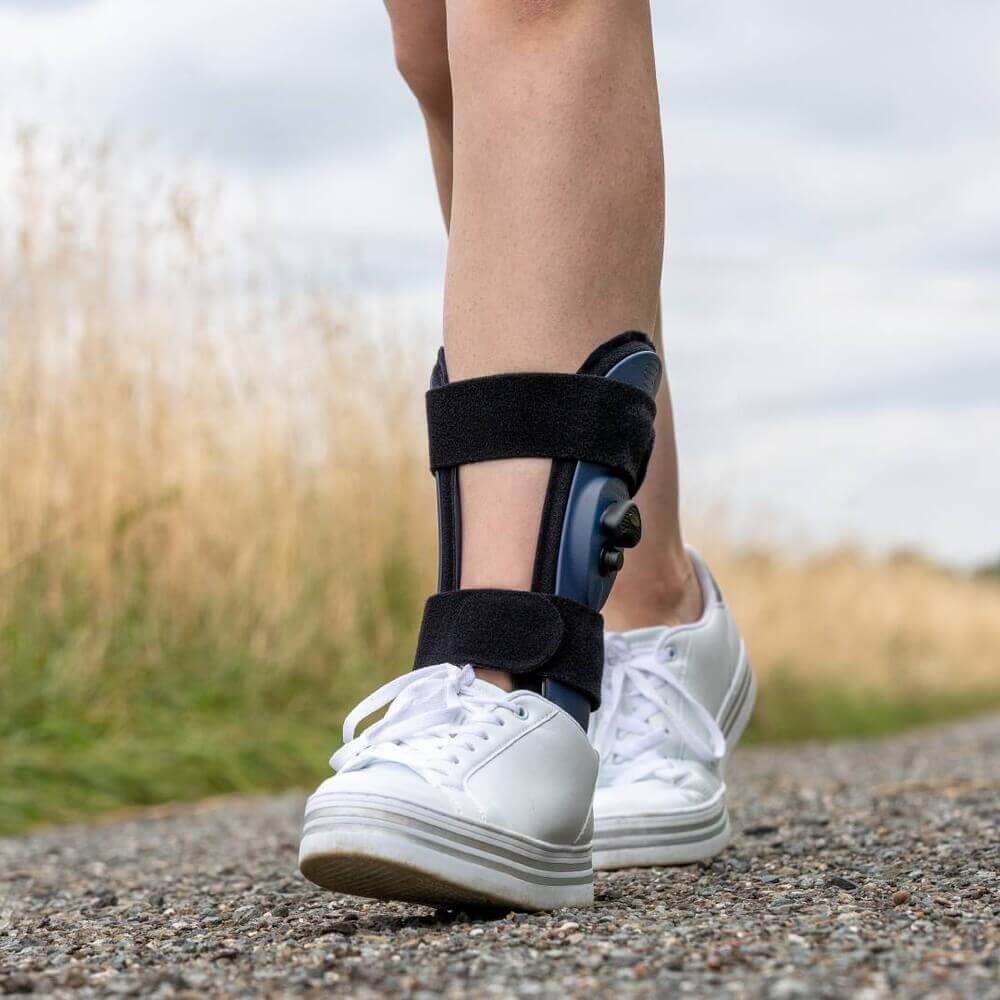 Detailaufnahme der Beine einer Person mit weissen Turnschuhen auf einem Kiesweg. Am linken Bein trägt die Person eine schwarze Fussorthese mit zwei Klettverschlüssen.