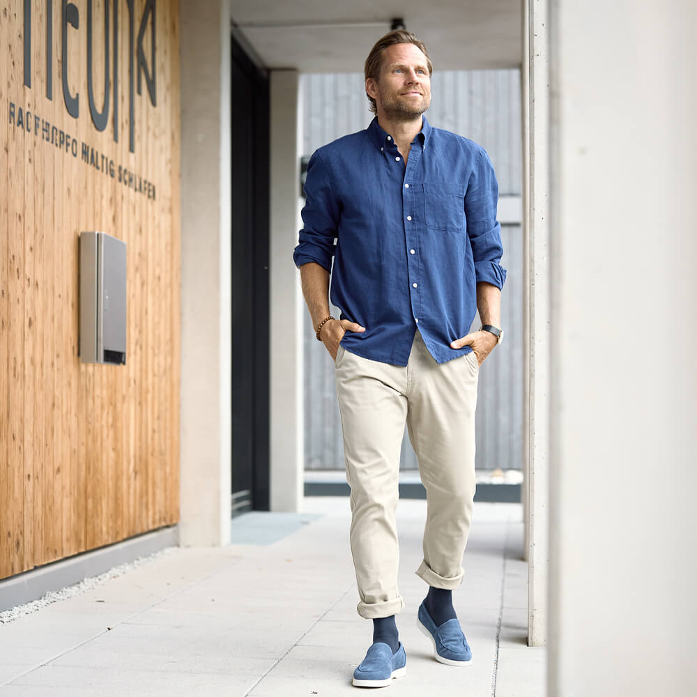 Mann in blauem Hemd und heller Hose geht mit den Händen in den Hosentaschen durch einen modernen Gebäudegang.
