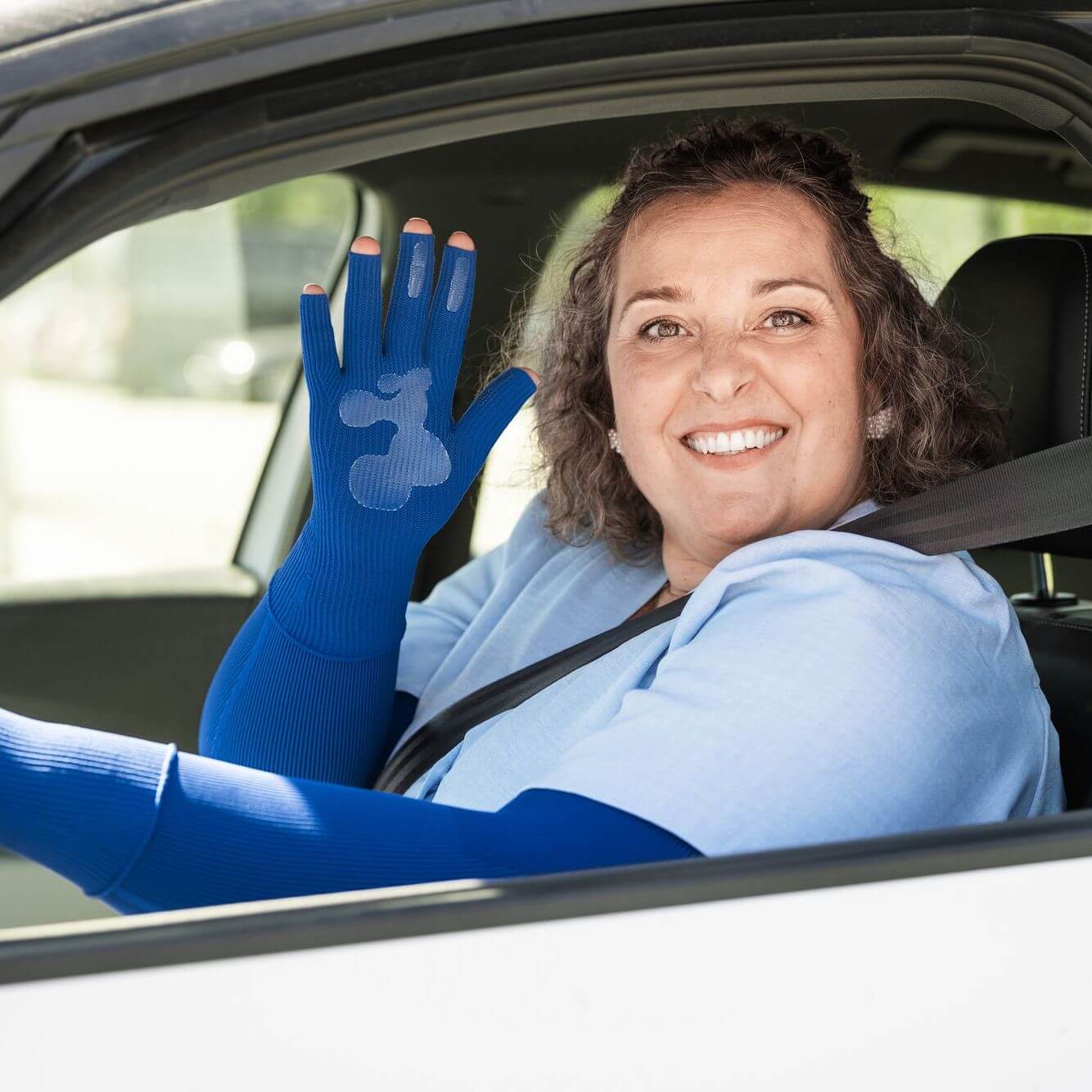 Lächelnde Frau mit Sicherheitsgurt im Auto, die einen blauen fingerlosen Kompressionshandschuh trägt und ihre Hand zeigt.