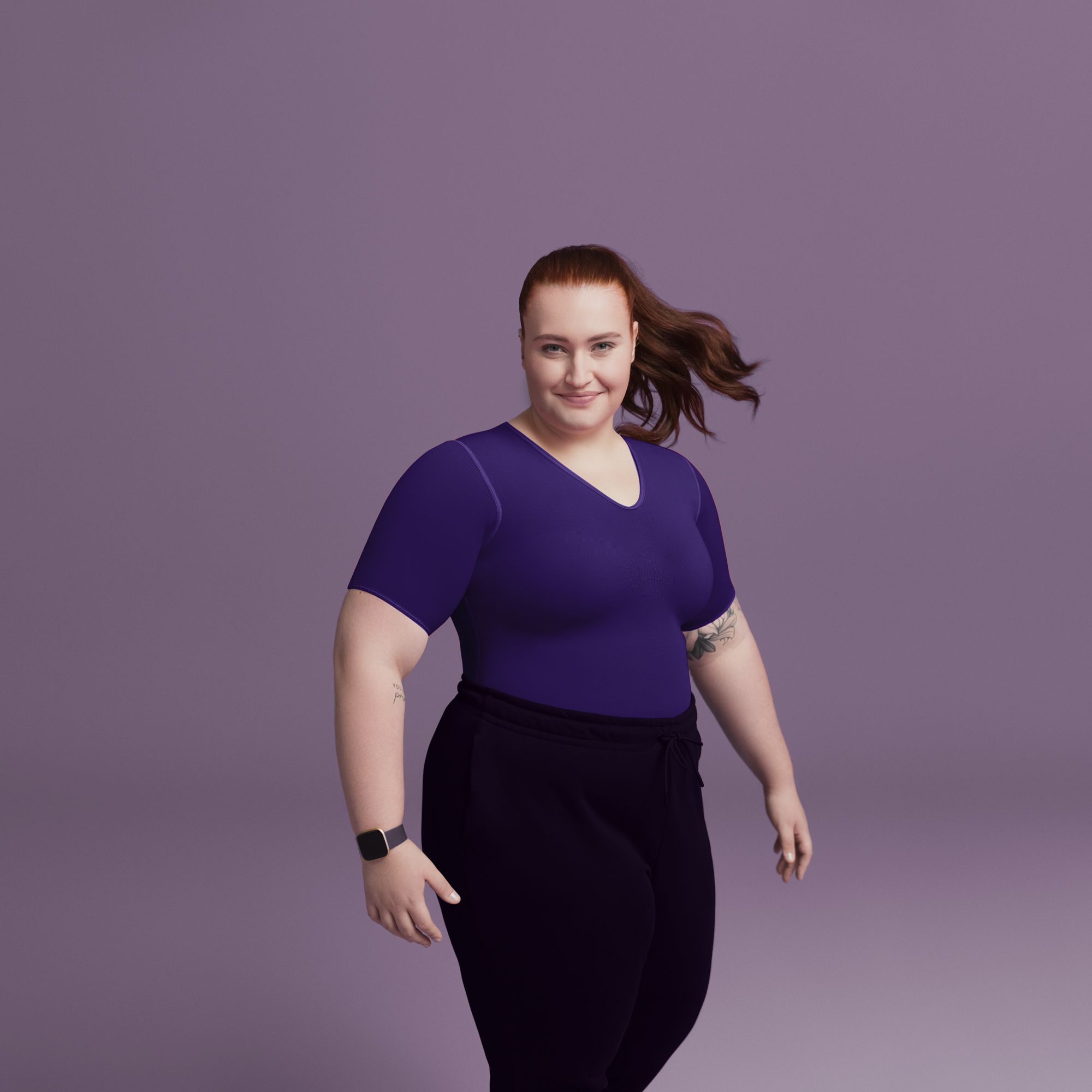 Frau mit langen, im Wind wehenden braunen Haaren steht vor einem einfarbigen lila Hintergrund. Sie trägt ein eng anliegendes, violettes T-Shirt, eine dunkle Hose mit Bindegürtel und eine Smartwatch am linken Handgelenk. Auf dem rechten Arm sind Tattoos sichtbar, und sie lächelt selbstbewusst in die Kamera.