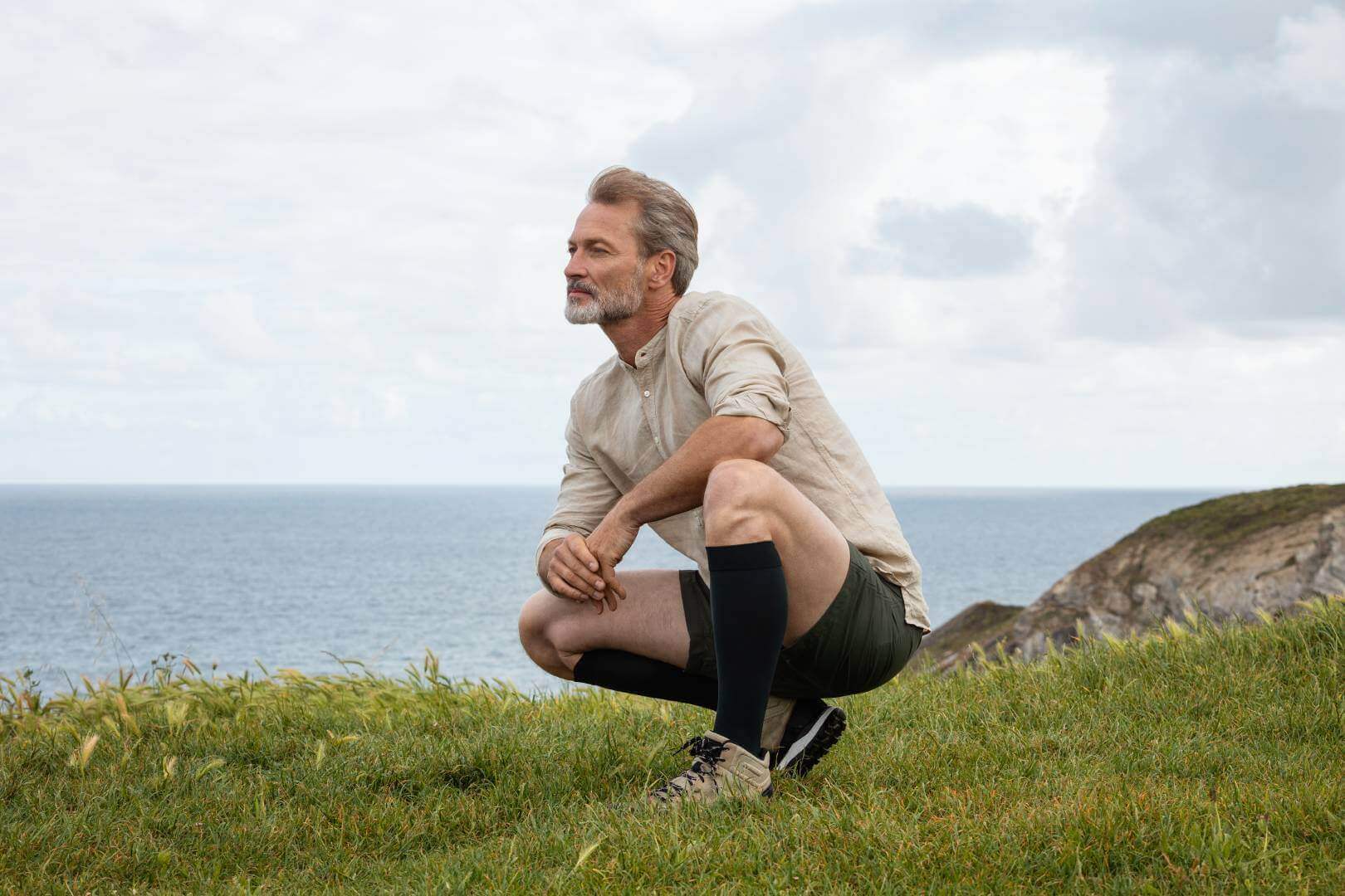 Ein Mann hockt auf einer Wiese am Meer. Er trägt Kompressionsstrümpfe von Bauerfeind.