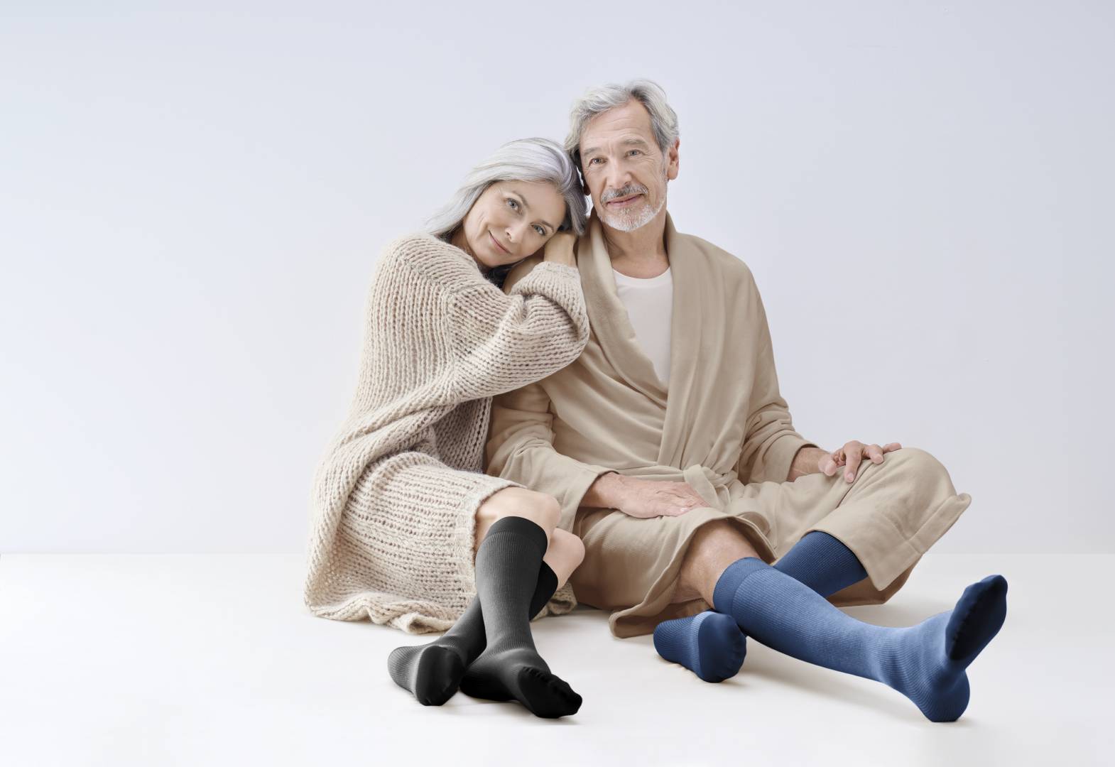 Ein älteres Paar sitzt entspannt nebeneinander. Beide tragen hautpflegende Kompressionsstrümpfe VenoTrain cocoon in verschiedenen Farben. Die Frau lehnt ihren Kopf liebevoll an die Schulter des Mannes.