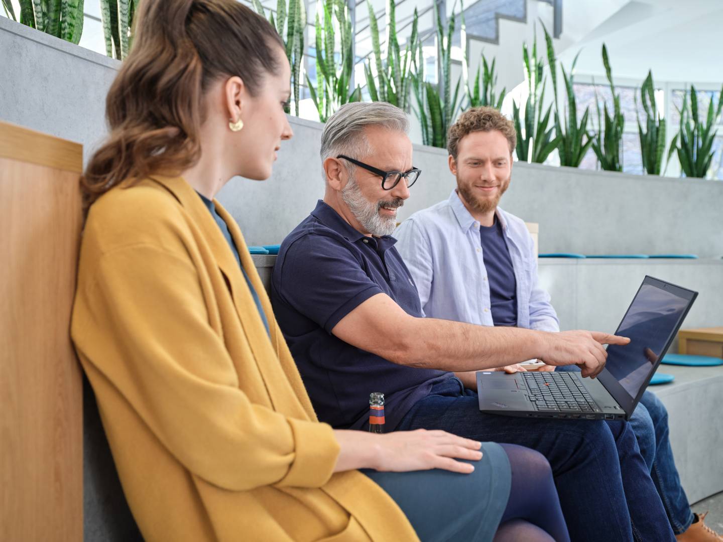 Drei Personen sitzen in einer modernen, begrünten Arbeitsumgebung. Ein Mann zeigt auf den Bildschirm eines Laptops, während die anderen aufmerksam zuhören. Die Szene vermittelt eine entspannte und produktive Atmosphäre.